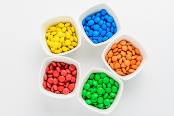 Five squared bowls with small red, yellow, blue, green and orange coated chocolate candies similar...