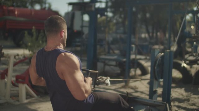 Rear view of muscular athletic fit man making low cable pulley row seated while training in outdoor gym. Strong concentrated bodybuilder working out on seated low row machine and flexing muscles.