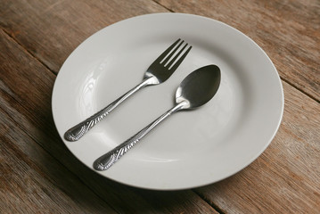 A white plate with spoon and fork on wooden background.