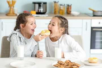 Portrait of cute twin girls eating donuts with milk in kitchen