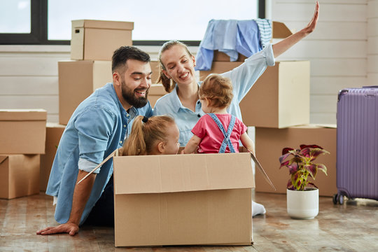 Caucasian Family, Man, Woman And Two Girls Play With Moving Box. Into It Sit Girls. Mother Keep Her Hand Like Airplane