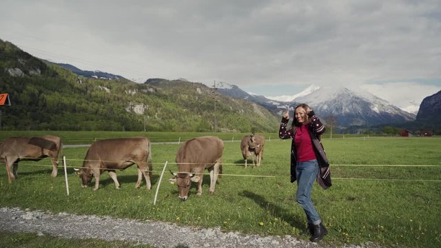 Beautiful young woman stands next to alpine cows in a pasture and smiling, feels happy, makes horns on her head with fingers