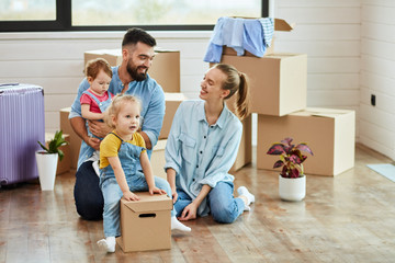 Fototapeta na wymiar Family of four men sit on floor in guest room in just sell house. Eldest daughter sit on box like rider. Mom and dad smile. Dad keep youngest daughter. Background moving boxes, suit and flower in pot