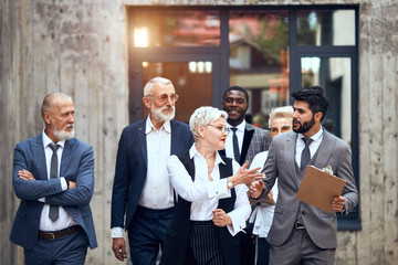 Five caucasian and one african businessmen wear different office suits move and discuss project outdoor. background big window