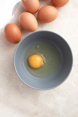 Raw uncooked egg in gray bowl and brown whole eggs on kitchen table, top view. Yellow egg yolk and liquid egg whites. Ingredient for baking, scrambled eggs or omelet