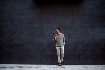 Handsome fashionable man smiling, leaning on gray wall and holding smart phone while looking away.