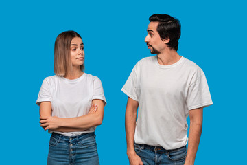 Fototapeta premium Couple of a young blond woman and brunet bearded man with mustaches in white t-shirts and blue jeans looking at each other posing isolated over blue background. Concept of an ideal couple.