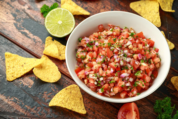 Mexican Tomato Salsa in white bowl with lime, red onion, jalapeno pepper, parsley and tortilla chips on wooden table