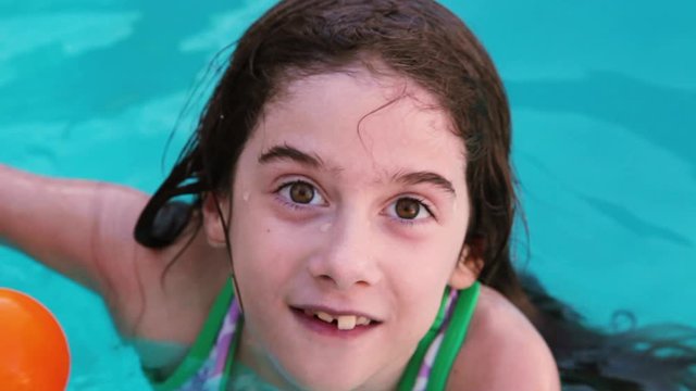 Looking down at a young girl with crooked teeth, thick eyebrows, and a very expressive face as she talks from inside the swimming pool. She is excited and it shows in her expressions. Wide eyes, mouth