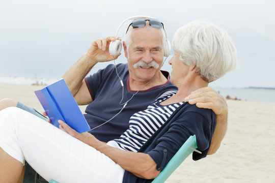 senior couple on beach reading book and listening to headphones - Powered by Adobe