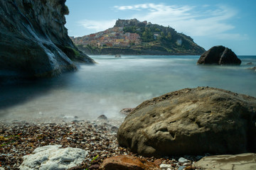 Fototapeta na wymiar castelsardo italy on the background with a milky blurry sea by long exposure. rocks in the front
