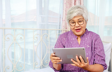 Portrait of smiling senior woman with eyeglasses using electronic tablet while relaxing at the bed room.