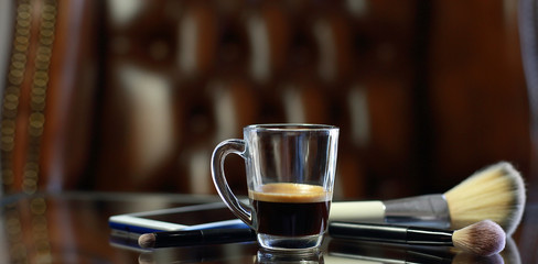 Cup of coffee with the phone and make-up set and brushes on the glass table