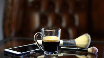 Cup of coffee with the phone and make-up set and brushes on the glass table