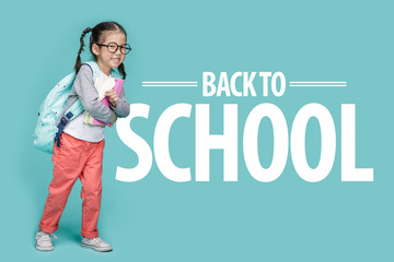 Beautiful smiling Asian little girl with glasses and hold a books with school bag is back to school, empty space in studio shot isolated on colorful blue background, Educational concept for school