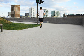 People, modern lifestyle, sports and wellness concept. Back view of sporty fit guy in running shoes exercising outdoors, doing cardio workout on embankment with multi storied buildings in background