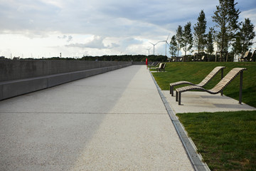 Outdoor summer image of broad river embankment with wooden desk-chairs or benches on green lawn along concrete promenade. Cloudy sky and wind turbines in background. Perfect urban location for walking