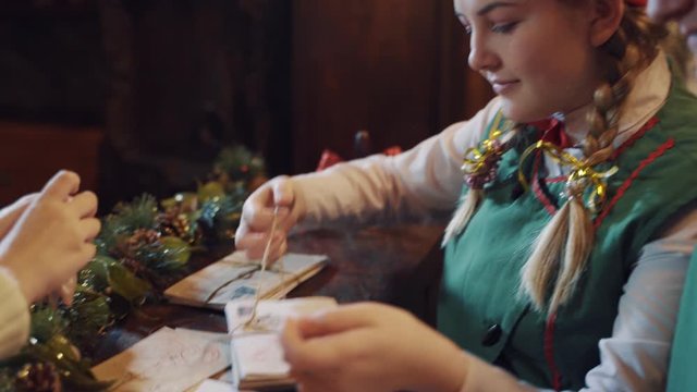 Elves and children making envelopes at Christmas. Many Christmas letters on the table and cute kid puts seal on the envelope.Female elf ties stack of envelopes.