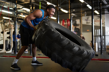 Full length portrait of muscular African man flipping tire during strength workout in modern gym, copy space