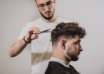 Young man with trendy haircut at barber shop. Barber does the hairstyle and beard trim.