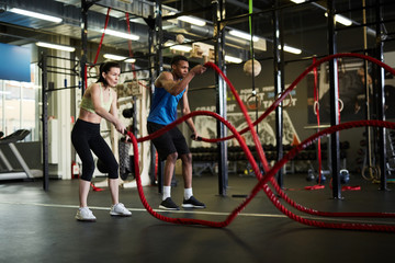 Side view portrait of fit couple exercising with battle ropes during strength workout in modern gym, copy space