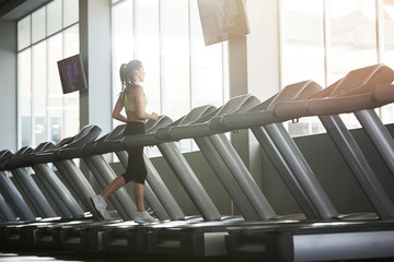 Wide angle back view portrait of young woman running on treadmill alone in empty gym, copy space