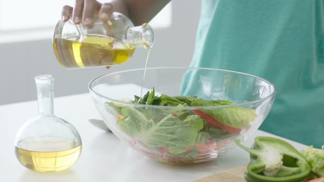 Woman preparing fresh salad in glass salad bowl, pouring oil over lettuce leaves and mixing with salad servers