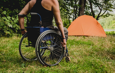 Disabled man resting in a campsite with friends. Wheelchair in the forest on the background of tents