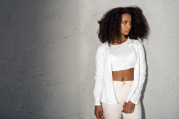 Freestyle. African girl in white outfit with bare belly standing isolated on gray looking aside...