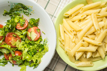 Plate with tubular pasta next to plate of tomato and cucumber salad.