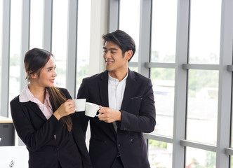 Obraz premium Couple of happy Asian colleagues talking while standing at the office window with cups of coffee.Concepts about teamwork, work fun and love