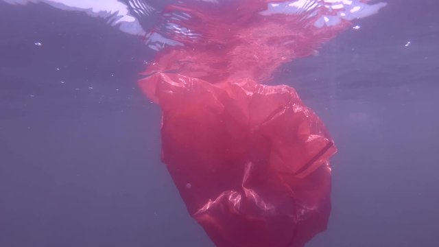Plastic pollution, a discarded rred plastic bag drifting in the blue water underw surface of water. Underwater shot