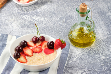 white bowl with oatmeal and strawberries and cherries and olive oil on a striped napkin and a gray background.