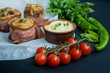Baked new potatoes wrapped in bacon, on top cheese, sauce and fresh vegetables. Dark background.