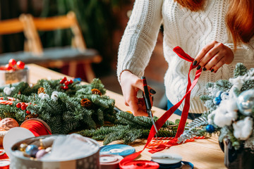Craft workshop. Cropped shot of female florist using red ribbon, green fir tree twig to create Christmas interior decoration.