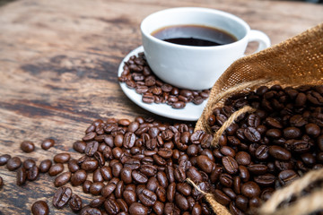 full of coffee beans spilling out bag on brown wooden background with a cup of black coffee