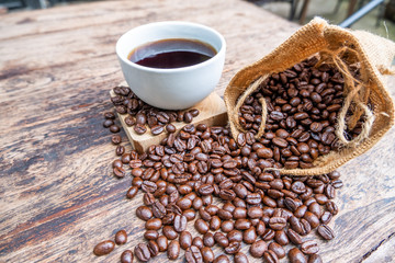 full of coffee beans spilling out bag on brown wooden background with a cup of black coffee