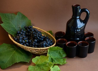  Still life of blue grapes in a wicker plate and wine in a clay jug with glasses on a brown background