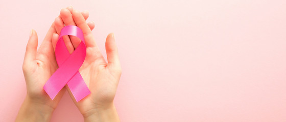 Breast cancer awareness campaign horizontal banner with copy space. Woman’s hands holding pink breast cancer awareness ribbon over pink background. Healthcare and medicine concept.