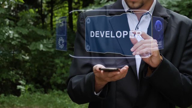 Unrecognizable businessman activates conceptual HUD holograms on smartphone with text Developer. Bearded man in a white shirt and a jacket with a holographic screen on a background of green trees