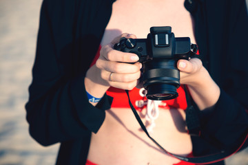 Girl holding modern mirrorless camera in her hands. Hobby and technology concept. Girl stands on the beach and looks at the digital camera.