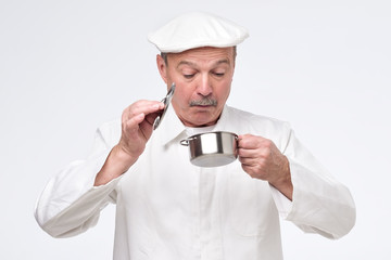 Senior chef cook holding small pan and looking inside it with surprise. Studio shot