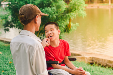 Fototapeta premium Disabled child on wheelchair and his dad, Sitting by the pond in the city park of Thailand, Sun set in the summer season, Life in the education age of special Children, Happy disabled kid concept.