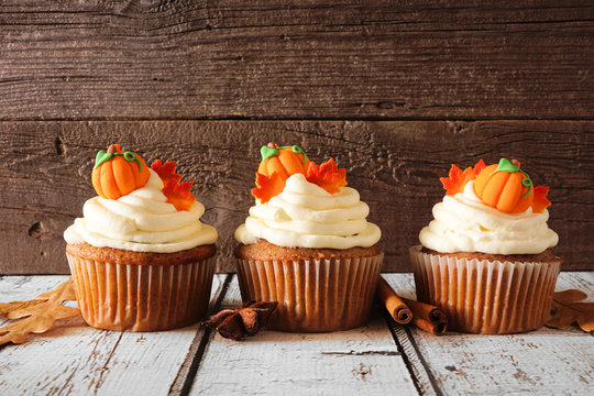 Fall pumpkin spice cupcakes with creamy frosting and autumn toppings. Row against a rustic wood background.