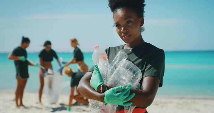 Slow motion of an young african female volunteer is smiling in camera satisfied with picking up a plastic litter on a beach with a sea to protect an environment.
