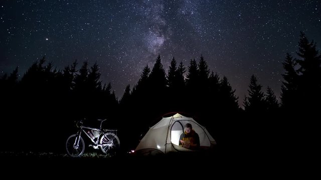 A Man Reads A Book In A Tent. The Milky Way Moves In The Night Sky Over The Silhouettes Of Trees. The Video Is Made Of Two Different Videos. 4K