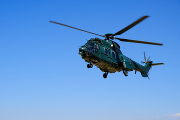 Big dark green hovering military helicopter on blue sky background