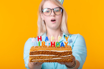 Portrait of a funny positive girl with a paper cap and glasses holding a congratulatory cake in her hands on a yellow background. Concept and fun and celebration. Advertising space.