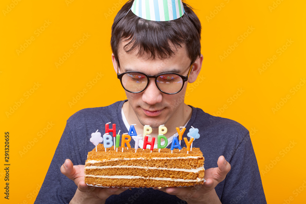 Wall mural Close-up of funny positive guy in glasses holds in his hands a homemade cake with the inscription happy birthday posing on a yellow background. Concept of holidays and anniversaries.