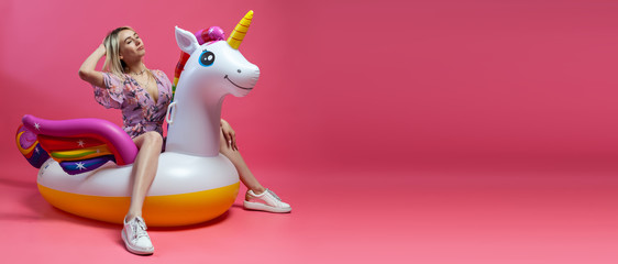 A beautiful blonde girl in a sexy sundress with slim legs in white sneakers sits on an inflatable multi-colored unicorn on a pink isolated background. Summer vacation at the beach and travel. © Aleksandr Kondratov